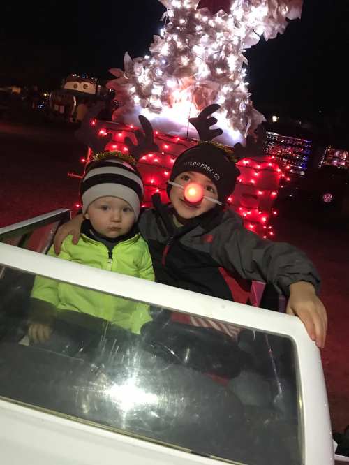 Two children sit in a toy car, surrounded by festive holiday lights and decorations at night.
