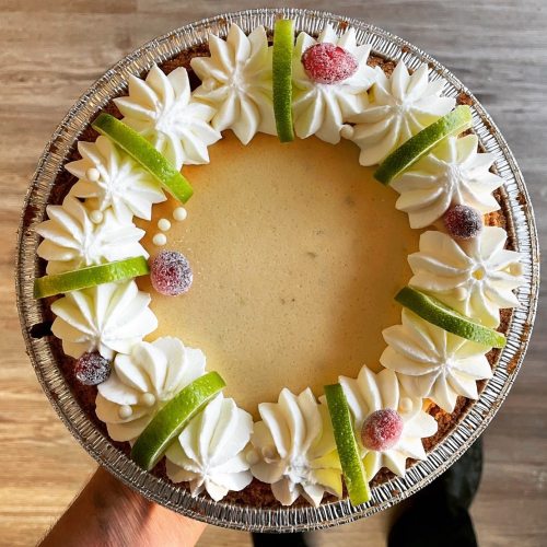 A round cheesecake topped with whipped cream, green apple slices, and red fruit garnishes, presented in a silver pie dish.