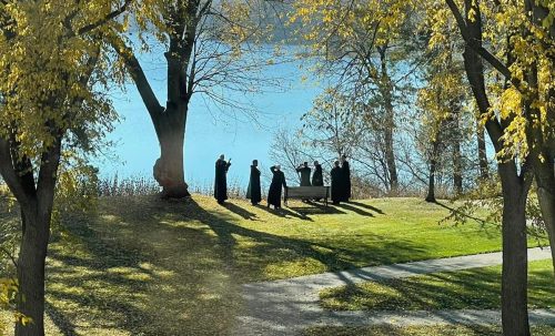 A group of people stands by a lake, surrounded by trees with vibrant yellow leaves on a sunny day.