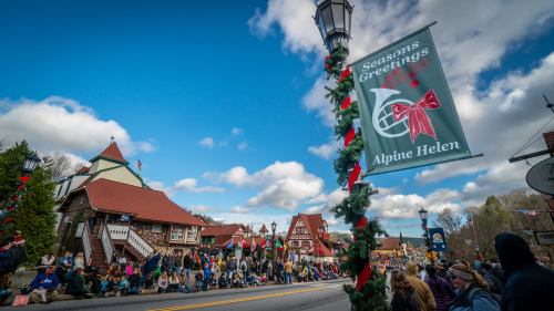 A festive street scene in Alpine Helen, with a holiday banner and crowds gathered for a parade under a blue sky.