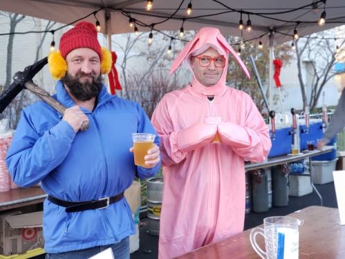 Two men stand under a tent, one in a blue jacket with a pickaxe, the other in a pink bunny costume, both holding drinks.