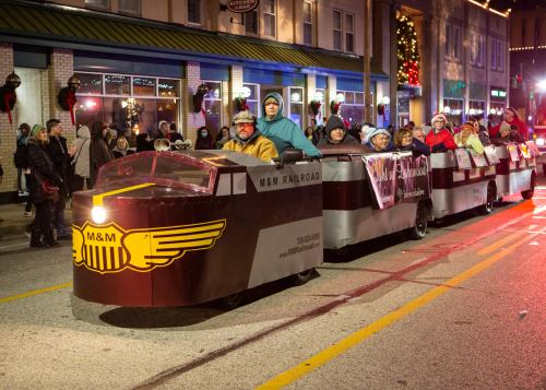 A festive parade float resembling a train, with people dressed warmly, celebrating in a decorated street at night.