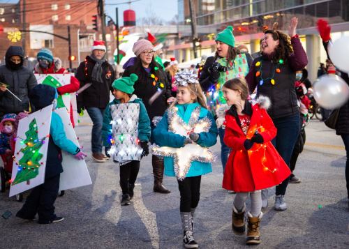 A festive parade scene with children and adults in holiday attire, holding decorations and smiling joyfully.