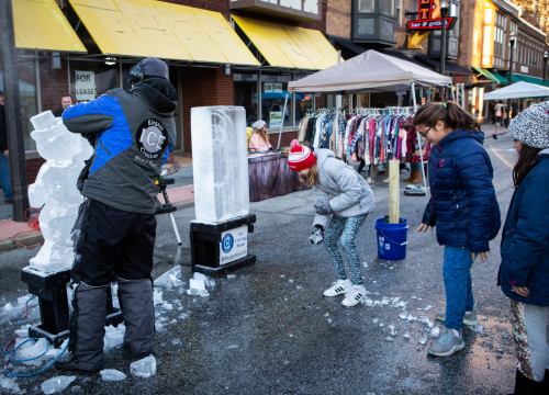 A person carves ice while children watch and play in a street market with clothing for sale in the background.