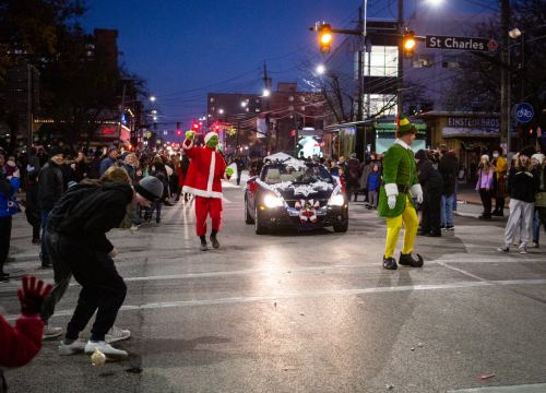 A festive parade scene with Santa, an elf, and a decorated car, surrounded by cheering crowds at dusk.