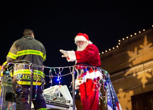 Santa Claus waves from a decorated fire truck, accompanied by a firefighter, with festive lights in the background.