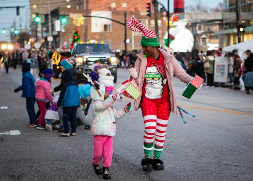 A festive parade scene with a woman in a Santa outfit and children holding gifts, surrounded by holiday decorations.