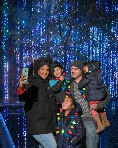A family of five takes a selfie in front of colorful holiday lights, smiling and wearing festive outfits.