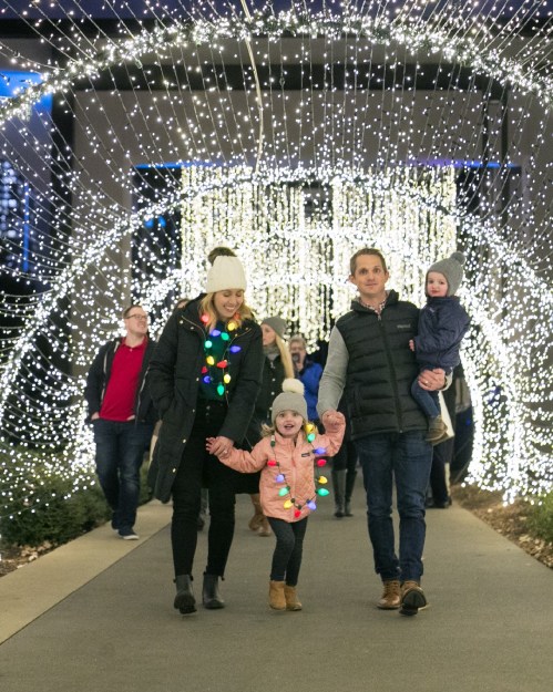 A family walks through a festive light display, with children wearing colorful lights and winter attire.