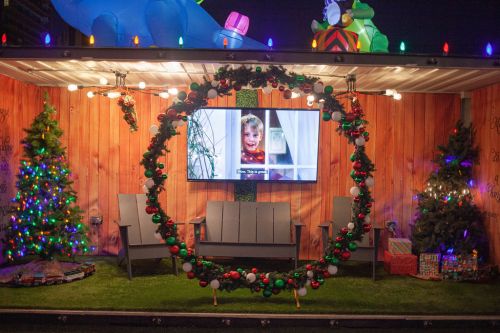 A festive outdoor scene with a decorated container, Christmas trees, lights, and a TV displaying a child's photo.