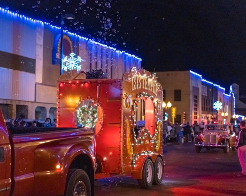 A festive parade scene at night with decorated floats, colorful lights, and bubbles floating in the air.