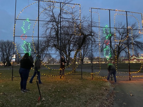 People setting up holiday lights in a park at dusk, with trees and festive decorations in the background.