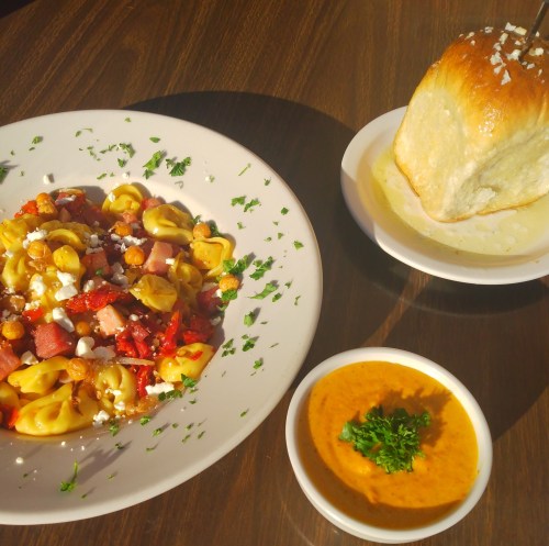 A plate of tortellini salad with ham, sun-dried tomatoes, and feta, alongside a bowl of soup and a bread roll.