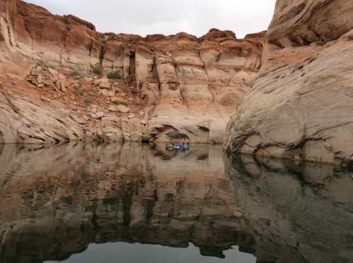 A serene landscape featuring towering red rock formations reflected in calm water, with a small boat in the distance.