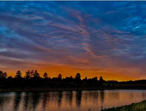 A serene lake at sunset, reflecting vibrant orange and blue skies with silhouettes of trees along the shore.