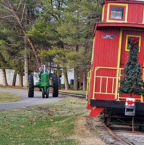 A green tractor drives past a red train car decorated with a Christmas tree, surrounded by trees and grass.
