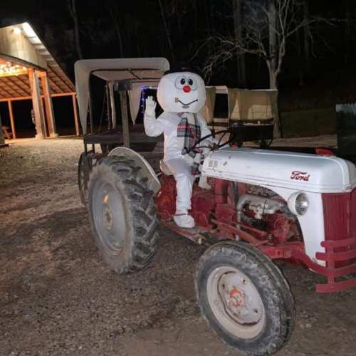 A person in a snowman costume sits on a vintage Ford tractor at night, with a barn in the background.