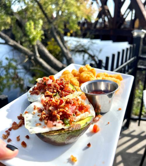 A plate of avocado halves topped with bacon and pico de gallo, served with tater tots and dipping sauce by a river.