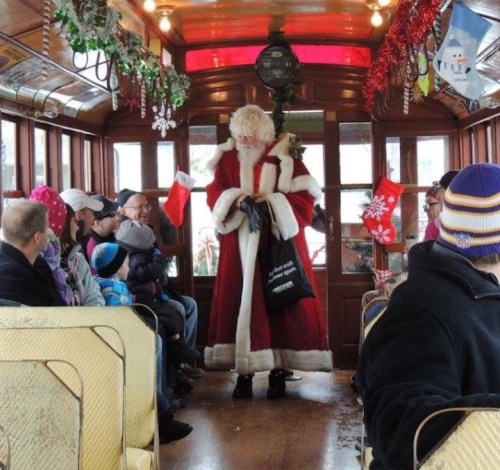 A festive trolley interior with passengers, as Santa in a red suit greets them, surrounded by holiday decorations.