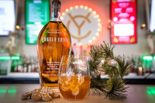 A bottle of Angel's Envy bourbon next to a glass of whiskey, gingerbread cookies, and a sprig of pine.