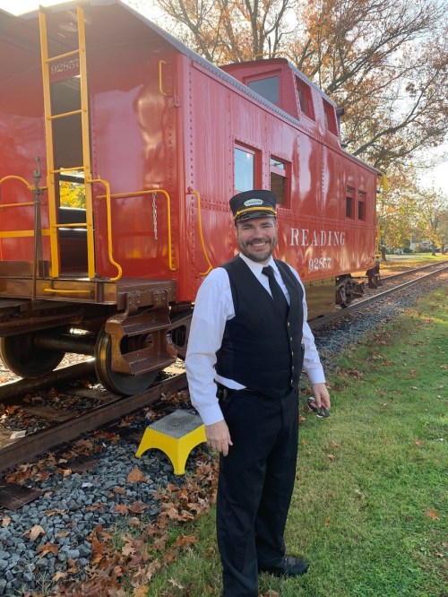 A smiling train conductor in a black vest and hat stands beside a red caboose on a sunny day.