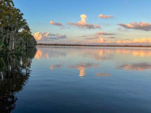A serene lake at sunset, with colorful clouds reflecting on the water's surface and trees lining the shore.