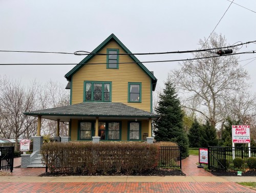A yellow house with green trim, surrounded by shrubs and trees, with a for sale sign in the front yard.