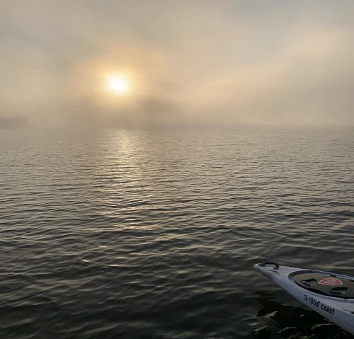 A serene scene of a foggy lake at sunrise, with soft light reflecting on the water and a kayak in the foreground.