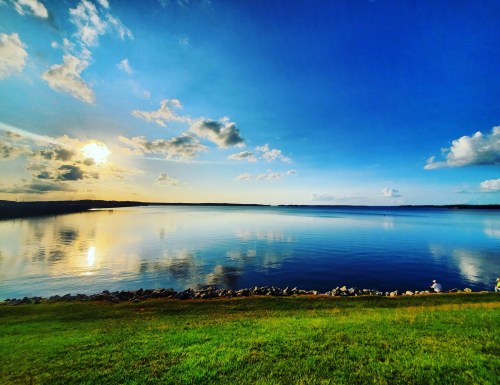A serene lake at sunset, with calm waters reflecting the sky and a grassy shore in the foreground.