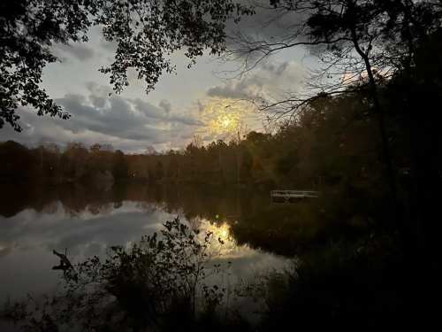 A serene lake at dusk, reflecting clouds and trees, with a hint of moonlight illuminating the water.