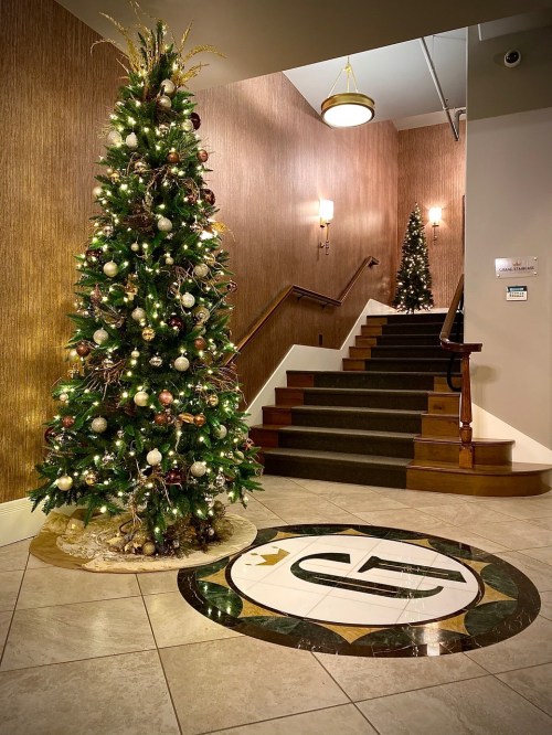 A decorated Christmas tree stands beside a staircase, with elegant wall decor and a circular logo on the floor.