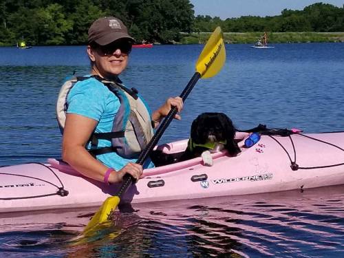 A woman in a pink kayak paddles on a calm lake with a dog beside her, surrounded by greenery and other kayakers.