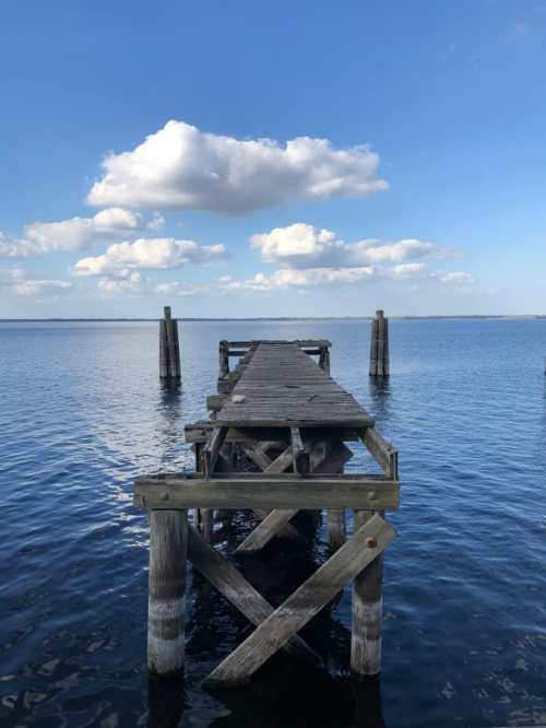 A weathered wooden pier extends into calm water under a blue sky with fluffy clouds.