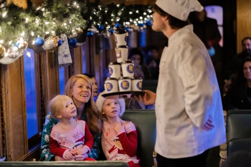 A chef presents a stack of cups to a smiling woman and two children on a festive train decorated with lights.