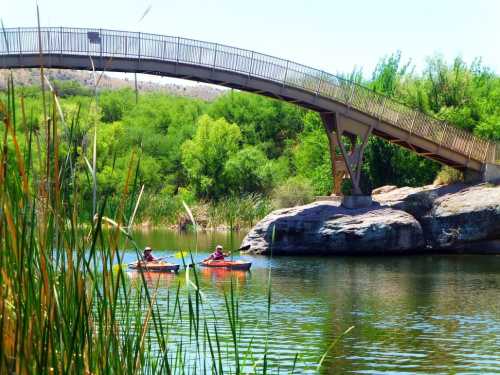 Two kayakers paddle on a calm river beneath a curved bridge, surrounded by lush greenery and rocky outcrops.