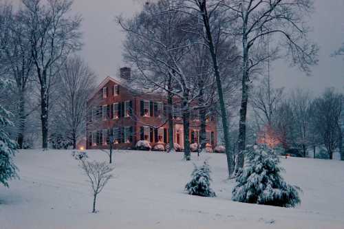 A brick house surrounded by snow-covered trees, softly lit in the evening glow of winter.