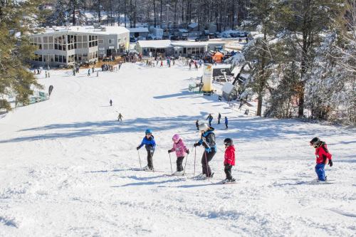 A snowy ski slope with children and adults skiing, surrounded by trees and a lodge in the background.