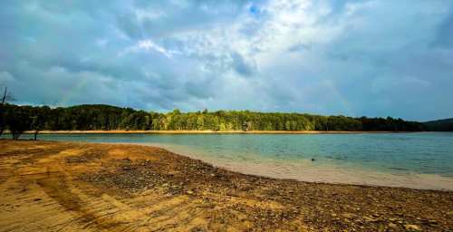 A serene lakeside view with a sandy shore, surrounded by lush green trees under a cloudy sky.
