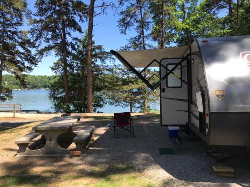 A camper parked by a lake, with a picnic table and chair under a tree-filled landscape.