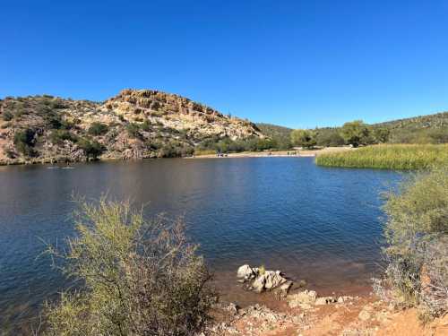 A serene lake surrounded by hills and greenery under a clear blue sky, with people enjoying the outdoors in the distance.