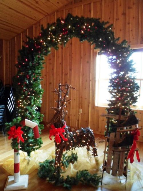 A festive arch made of greenery and lights, featuring a reindeer and sled, set in a wooden room.