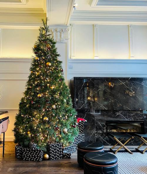 A beautifully decorated Christmas tree with gold ornaments and wrapped gifts, beside a sleek black marble fireplace.