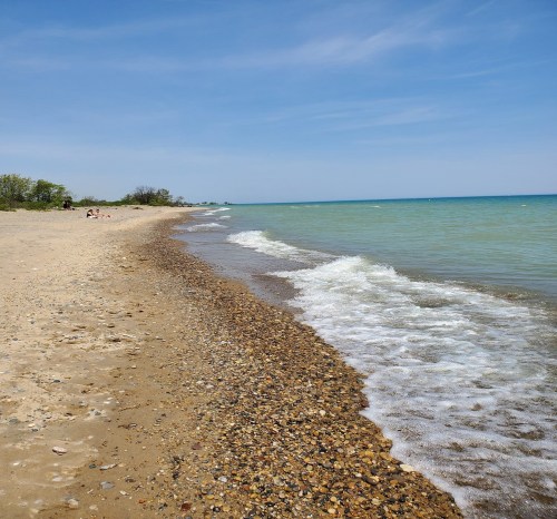 A sandy beach with pebbles, gentle waves lapping at the shore, and a clear blue sky in the background.
