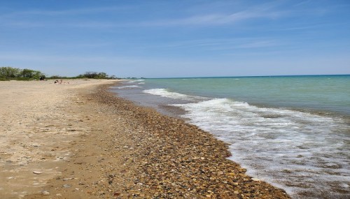 A sandy beach with pebbles, gentle waves lapping at the shore, and a clear blue sky in the background.