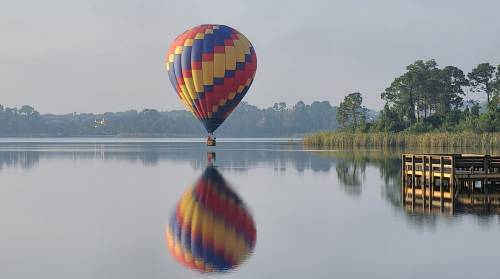 A colorful hot air balloon floats above a calm lake, reflecting in the water, surrounded by trees and a wooden dock.