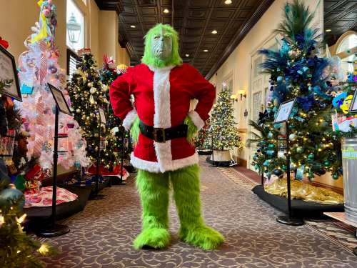 A green, furry character in a Santa suit stands confidently among decorated Christmas trees.