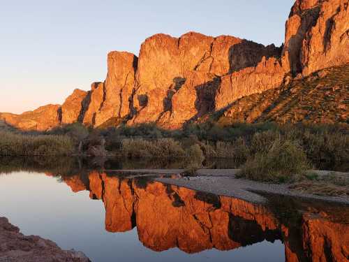 Sunset over rugged mountains reflecting in a calm river, surrounded by lush greenery.