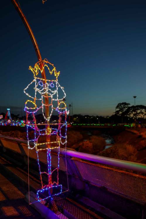 A colorful light display of a nutcracker figure on a bridge at dusk, with festive decorations in the background.