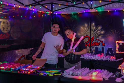 Two people at a festive booth with colorful lights, showcasing glow sticks and other light-up items.