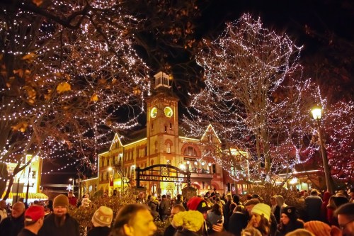 A festive crowd gathers at night, surrounded by twinkling holiday lights on trees and a historic building.